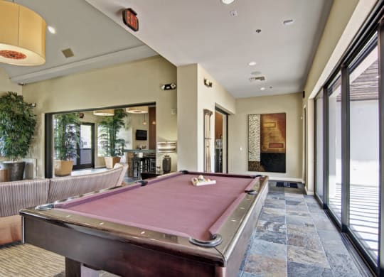 Modern recreation room here at Natura Villas Apartments with a burgundy pool table, abstract wall art, potted plants, large windows, and cozy seating. Inviting and relaxed atmosphere.