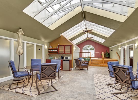 Bright sunroom here at The Club at Eagle Pointe Apartments with skylights, wicker furniture with blue cushions, patterned rugs, a kitchenette with a microwave, and a calming hot tub area.