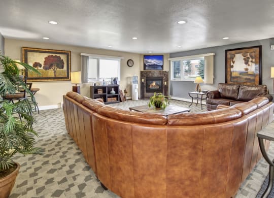 Cozy resident lounge here at The Club at Eagle Pointe Apartments with a large brown leather sectional, centered around a fireplace. Decor includes landscape paintings, a table lamp, and a potted plant.