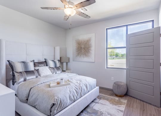 Bright bedroom here at 49 West Apartments with a large bed featuring gray and silver pillows and comforter, modern art on the wall, a ceiling fan, and a sunlit window. Cozy and serene atmosphere.
