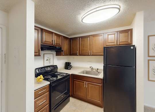 A bright, efficient kitchen here at Austin Park featuring rich wood cabinetry, black appliances including a refrigerator and electric range, light countertops, a stainless steel sink, and neutral flooring, with overhead lighting and a compact layout that offers practical storage and a clean, functional space for everyday cooking.