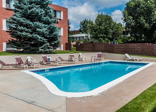 A refreshing outdoor swimming pool here at Bristol Square showcasing clear blue water, a curved concrete pool deck, red and white lounge chairs, green grass edging, mature trees, and brick apartment buildings under a bright sky with scattered clouds.