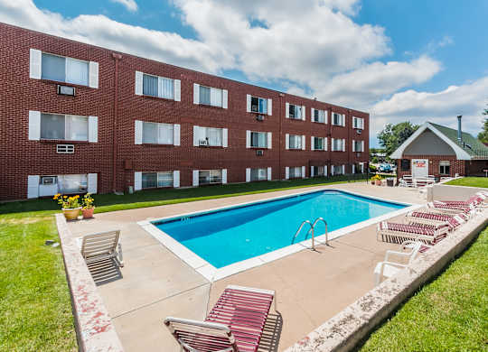 A sunny outdoor swimming pool here at Bristol Square featuring clear blue water, a rectangular pool with metal handrails, red and white lounge chairs on a concrete deck, trimmed green grass, and a brick apartment building backdrop beneath a bright sky with soft white clouds.