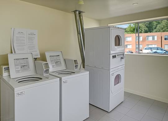 A clean shared laundry room here at Bristol Square featuring white top-load washing machines and stacked dryers, light tile flooring, neutral walls, posted laundry instructions above the machines, and a large window providing natural light and views of the apartment community outside.