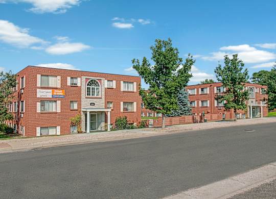 A charming brick apartment community exterior here at Bristol Square featuring multiple three-story residential buildings with white shutters, arched entryway details, mature trees, manicured landscaping, and a quiet street view under a bright blue sky.