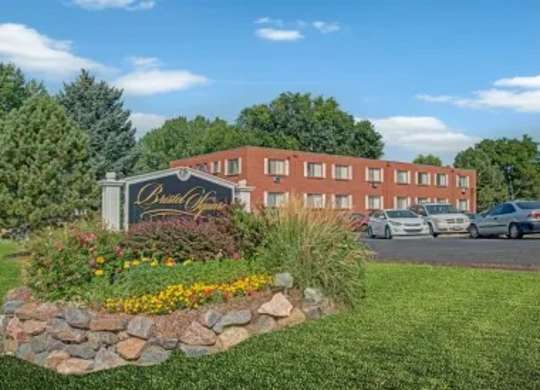 A welcoming apartment community entrance sign here at Bristol Square showcasing a landscaped monument sign surrounded by colorful flowers, decorative stone edging, green lawn, mature trees, parked vehicles, and classic brick apartment buildings beneath a bright blue sky.