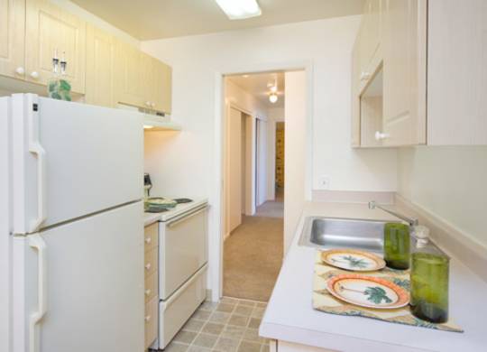 A clean and functional kitchen here at Bristol Square showcasing light wood cabinetry, white appliances including a full-size refrigerator and range, laminate countertops with a stainless steel sink, decorative place settings, and a tiled floor that connects to a carpeted hallway, creating a bright and practical cooking space.