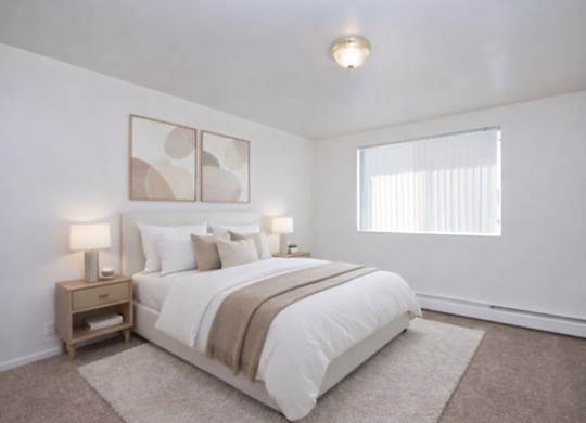 A calm and inviting bedroom here at Bristol Square featuring a queen-sized bed with crisp white bedding and a taupe throw blanket, matching wood nightstands with modern lamps, soft neutral wall art above the headboard, plush carpet flooring, and a wide window with vertical blinds that brings in gentle natural light.