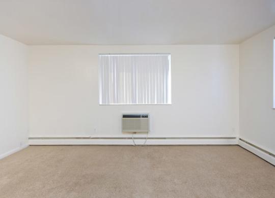A bright and open living room here at Bristol Square featuring plush carpet flooring, soft neutral walls, a wide window with vertical blinds, and a wall-mounted air conditioning unit beneath the window, offering a clean layout with ample natural light and flexible space for comfortable living and furniture placement.