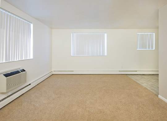 A spacious and sunlit living room here at Bristol Square showcasing soft neutral walls, plush carpet flooring, multiple wide windows with vertical blinds, a wall-mounted air conditioning unit, and an open layout that transitions into an adjacent dining area with durable flooring, creating a bright and versatile living space.