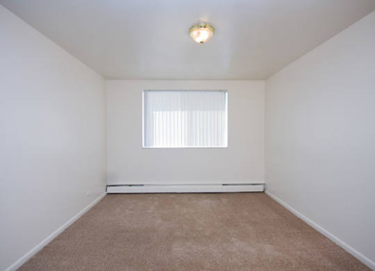 A bright and cozy bedroom here at Bristol Square featuring soft neutral walls, plush carpet flooring, a wide window with vertical blinds that allows natural light to fill the room, a simple ceiling light fixture, and a clean rectangular layout that offers flexibility for bedroom furniture and comfortable everyday living.