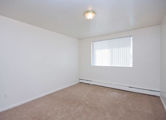 A bright and airy bedroom here at Bristol Square featuring soft white walls, plush carpet flooring, a wide window with vertical blinds for natural light, baseboard heating along the wall, and a simple ceiling light fixture, creating a clean and comfortable space suitable for a variety of bedroom furniture layouts.