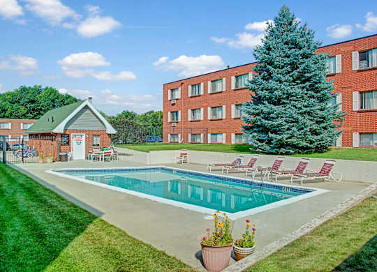 A sunny outdoor swimming pool here at Bristol Square featuring a rectangular pool surrounded by a concrete deck, red lounge chairs, green lawn landscaping, and a tall evergreen tree set against brick apartment buildings and a bright blue sky with scattered clouds.