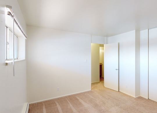 A bright bedroom here at Copper Stone featuring soft carpet flooring, clean white walls, and a spacious layout with a wide closet with sliding doors, a window allowing natural light, and an open doorway leading to the adjacent bathroom, creating a simple and flexible space ready for personalization.
