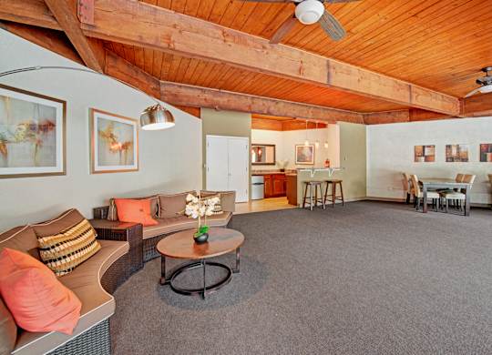 A warm, inviting community lounge here at Copper Stone featuring a wood plank ceiling with exposed beams and ceiling fans, cozy sectional seating with accent pillows, a round coffee table, dining tables and chairs, and a small bar area, all set within a spacious, carpeted interior designed for gathering and relaxation.