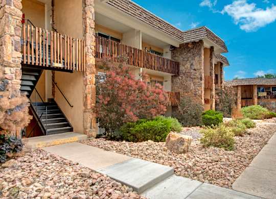 A landscaped exterior walkway here at Copper Stone featuring stone-accented apartment buildings with private wood-railed balconies, exterior stair access, desert-style rock landscaping, shrubs and flowering bushes, and a clean concrete path creating an inviting residential setting.