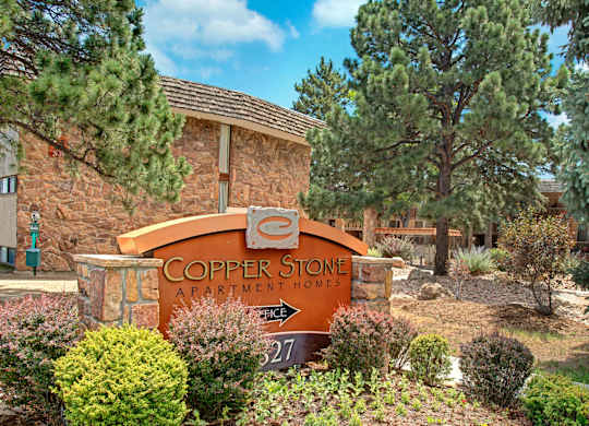 A welcoming entrance monument sign here at Copper Stone featuring a curved copper-toned panel with stone accents, surrounded by manicured shrubs, flowering plants, and mature pine trees, with rustic stone apartment buildings visible in the background under a bright blue sky.