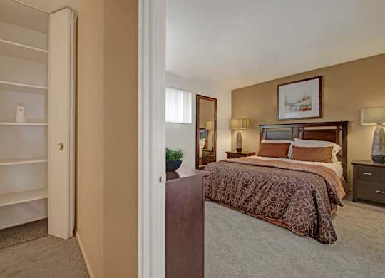 A warm bedroom here at Copper Stone showcasing a wood bed frame with patterned bedding, matching nightstands with lamps, soft carpet flooring, neutral wall tones, framed artwork above the headboard, a tall mirror, and an open shelving closet visible from the entry that adds functional storage and a comfortable, inviting layout.