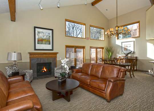A cozy living room here at Eagle Ridge featuring leather sofas arranged around a round coffee table, a fireplace with a wood mantel, and patterned armchairs, complemented by large windows that bring in natural light, vaulted ceilings with wood beams, and an adjacent dining area with a chandelier visible in the background.