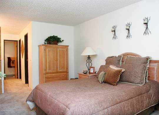 A cozy bedroom here at Eagle Ridge showcasing warm neutral tones with a wood bed frame dressed in textured bedding, matching wood nightstands with a table lamp, soft carpeted flooring, a tall wardrobe for added storage, and wall decor above the headboard, creating a comfortable and inviting retreat connected to a hallway leading toward the bathroom.