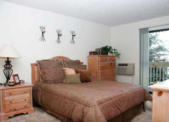 A cozy bedroom here at Eagle Ridge featuring a wood bed frame with layered neutral bedding, matching wood nightstand and dresser, soft carpeted flooring, decorative wall accents above the headboard, and a sliding glass door that brings in natural light and opens to a private balcony overlooking trees.
