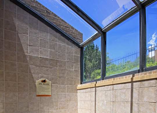A bright indoor spa area here at Eagle Ridge featuring a tiled hot tub with gently bubbling water, a built-in handrail for easy access, neutral tile walls and flooring, and large angled windows overhead that allow abundant natural light and views of greenery outside.