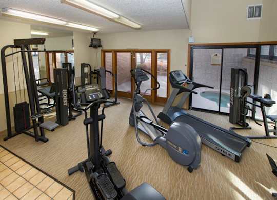 A bright fitness center here at Eagle Ridge showcasing a well-equipped workout space with treadmills, an elliptical, strength training machines, and free weights arranged on carpeted flooring, with large windows and glass doors providing natural light and views into the adjacent indoor pool area.
