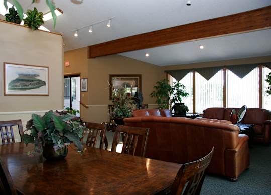 A welcoming community gathering space here at Eagle Ridge featuring a wooden dining table with matching chairs in the foreground, brown leather sofas arranged for conversation, exposed wood ceiling beams, neutral walls with framed artwork, tall windows with vertical blinds letting in natural light, and greenery accents that create a warm, comfortable atmosphere.