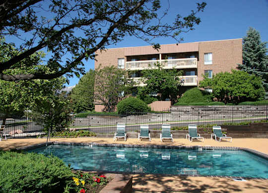 A sunlit outdoor pool area here at Eagle Ridge showcasing a clear swimming pool bordered by a spacious concrete deck with lounge chairs, mature trees providing shade, landscaped greenery and flowers in the foreground, and a multi-story brick apartment building with balconies rising in the background under a bright blue sky.