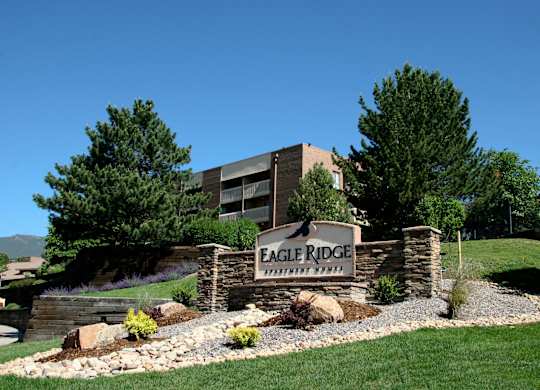 A welcoming stone monument sign here at Eagle Ridge set on a gently sloped landscaped hillside, featuring layered stonework, gravel accents, and low shrubs, with mature pine trees and a multi-story apartment building rising behind it under a clear blue sky.