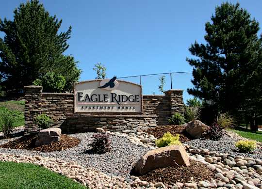 A prominent stone monument sign here at Eagle Ridge set within manicured landscaping, featuring layered stonework, decorative rock beds, low shrubs, and large accent boulders, framed by tall evergreen trees and a gently sloped grassy hillside beneath a clear blue sky.