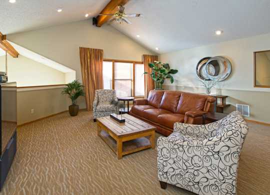 A spacious living room here at Eagle Ridge showcasing vaulted ceilings with exposed wood beams, a leather sofa centered around a wooden coffee table, patterned accent chairs, soft carpeted flooring, and large windows with warm curtains that bring in natural light and create an inviting, comfortable gathering space.