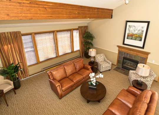 A bright, spacious living room here at Eagle Ridge featuring vaulted ceilings with exposed wood beams, warm neutral walls, leather sofas arranged around a round coffee table, patterned accent chairs, a cozy fireplace with a wood mantel, soft carpeted flooring, and large windows with blinds that allow natural light to fill the inviting space.