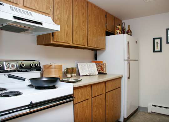 A cozy kitchen here at Eagle Ridge featuring warm wood cabinetry, light laminate countertops, a white refrigerator and range, upper and lower storage, neutral flooring, simple wall art, and staged countertop accents that highlight a practical layout designed for everyday cooking and convenience.