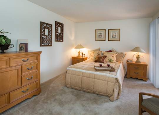 A comfortable bedroom here at Eagle Ridge featuring a neatly made bed with layered neutral bedding, matching wood nightstands with soft table lamps, framed wall art above the headboard, plush carpeted flooring, a spacious dresser for storage, and a sliding glass door that brings in natural light and connects the room to an outdoor area.