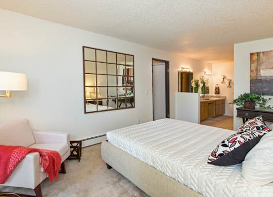 A bright bedroom here at Eagle Ridge showcasing a spacious sleeping area with a neatly made bed, plush carpeted flooring, a cozy upholstered accent chair with a floor lamp, decorative wall mirror, and an open layout that leads toward a vanity and bathroom area, creating a comfortable and functional retreat with warm lighting and clean finishes.