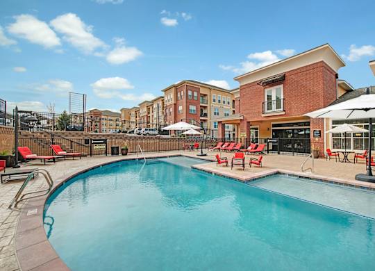 A resort-style outdoor pool here at Elements at Briargate featuring a curved swimming pool with a shallow entry, surrounding stone deck, red lounge chairs with umbrellas, fenced seating areas, and a clubhouse backdrop, all set against multi-story apartment buildings under a bright blue sky.