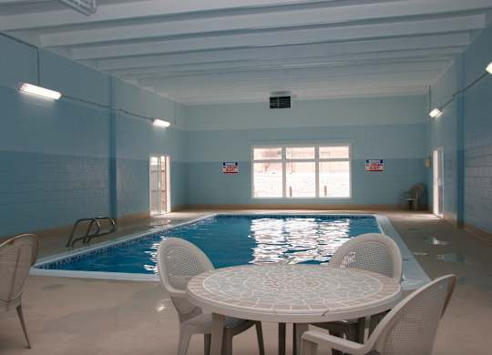 A spacious indoor pool area here at Hyde Park North Apartments with a rectangular swimming pool surrounded by a concrete deck. Natural light enters through large windows, while poolside tables and chairs offer space to relax. Light blue walls, overhead lighting, and easy pool access create a comfortable, year-round amenity.