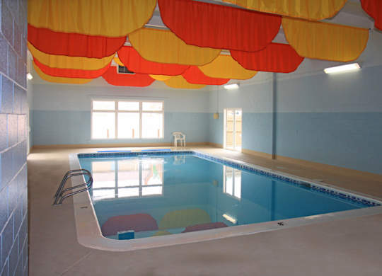 A bright indoor pool here at Hyde Park North Apartments featuring a rectangular swimming pool surrounded by a smooth concrete deck. Large windows allow natural light into the space, while colorful ceiling panels add visual interest. Pool steps and handrails provide easy access, creating a comfortable year-round amenity for residents.