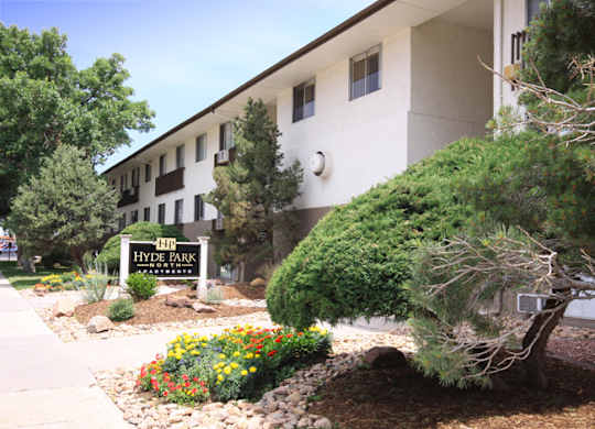 A landscaped community entrance here at Hyde Park North Apartments featuring a monument-style sign surrounded by colorful flowers, decorative rock beds, and mature trees. A multi-story apartment building with large windows sits behind the sign, creating a welcoming and well-maintained first impression of the community.