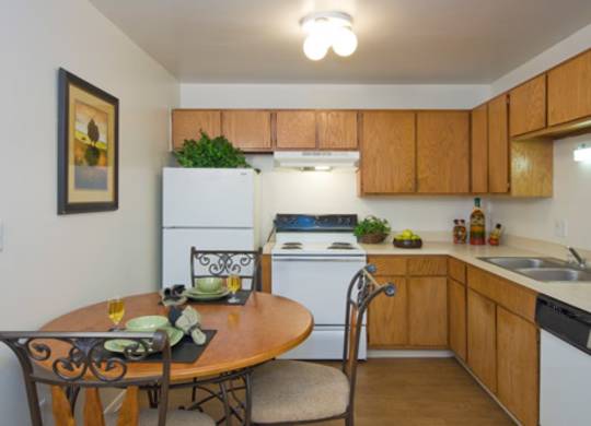 A welcoming kitchen here at Hyde Park North Apartments with warm wood cabinets, laminate countertops, and neutral flooring. A round dining table with seating sits beside the kitchen, while a refrigerator, stove, dishwasher, and sink are arranged in an efficient layout, creating a comfortable space for everyday cooking and dining.