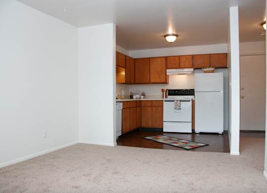 A bright, open living and kitchen area here at Hyde Park North Apartments with beige carpet in the living space and wood-style flooring in the kitchen. Warm wood cabinets, light countertops, a refrigerator, stove, dishwasher, and sink create a functional kitchen, while the open layout offers a smooth flow between living, dining, and cooking areas.