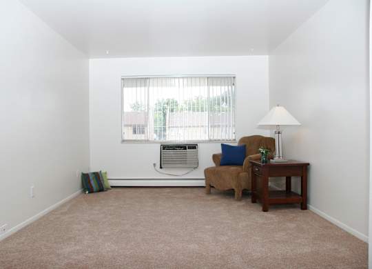 A spacious living room here at Hyde Park North Apartments with beige carpet and clean white walls, featuring a wide window with vertical blinds that brings in natural light. A wall-mounted climate unit sits below the window, while a cozy chair, side table, and lamp highlight the room’s simple layout and comfortable feel.