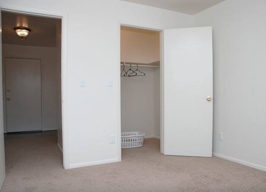 A bright bedroom here at Hyde Park North Apartments with beige carpet and clean white walls, featuring an open closet with hanging space and shelving for storage. A doorway leads to the hallway, while the simple layout offers flexibility for bedroom furniture and creates a comfortable, easy-to-personalize living space.