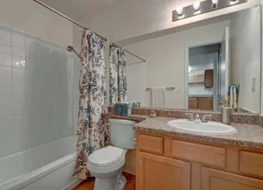 A clean, functional bathroom here at Hyde Park Apartments with a bathtub and shower combo surrounded by white tile, a white toilet, and a wooden vanity topped with a speckled countertop. A wide mirror with overhead lighting brightens the space, while neutral finishes create a simple and practical layout.