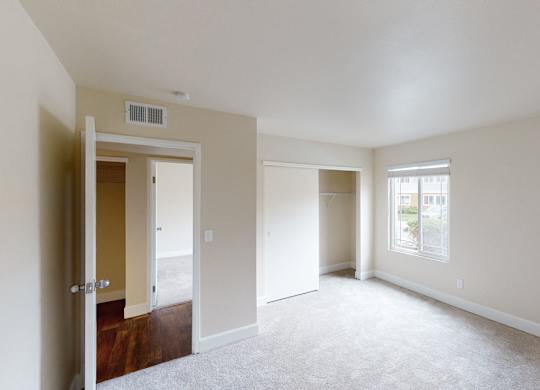 A bright neutral bedroom here at Retreat at Austin Bluffs featuring soft beige walls, plush carpet flooring, and a clean, open layout that feels spacious and inviting. A sliding-door closet with built-in shelving provides convenient storage, while a large window brings in natural light and views of the surrounding community.