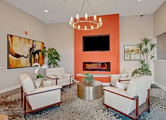 A stylish resident lounge here at Vista View featuring a bold accent wall with a built-in fireplace, a mounted television, modern upholstered seating arranged around a round coffee table, contemporary artwork, indoor plants, and a statement chandelier creating a warm and inviting community gathering space.