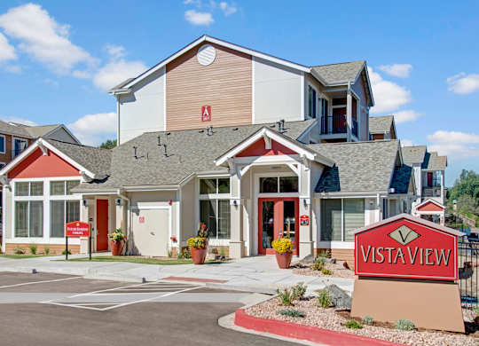 A welcoming exterior clubhouse building here at Vista View featuring peaked rooflines, neutral siding with red accent panels, large windows, and a clearly marked entrance, with landscaped planters, paved walkways, and a prominent Vista View monument sign set against a bright blue sky with scattered clouds.