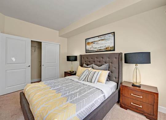 A cozy staged bedroom here at Vista View featuring a tufted upholstered bed with patterned neutral bedding, matching wood nightstands with modern lamps, soft carpet flooring, and sliding closet doors, complemented by warm wall tones and simple artwork that create a comfortable and inviting sleeping space.