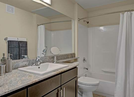 A bright bathroom here at Vista View showcasing a clean vanity with a stone-look countertop and integrated sink, a large wall mirror that reflects the space, a toilet beside a tub and shower combination with a curved shower rod and white curtain, and neutral wall tones that create a fresh, functional, and well-lit bathroom layout.